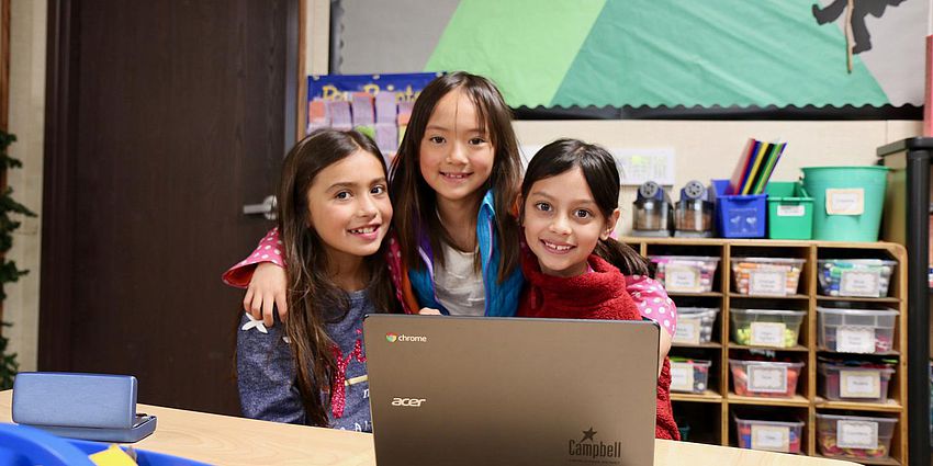 three girls hugging in front of a chromebook