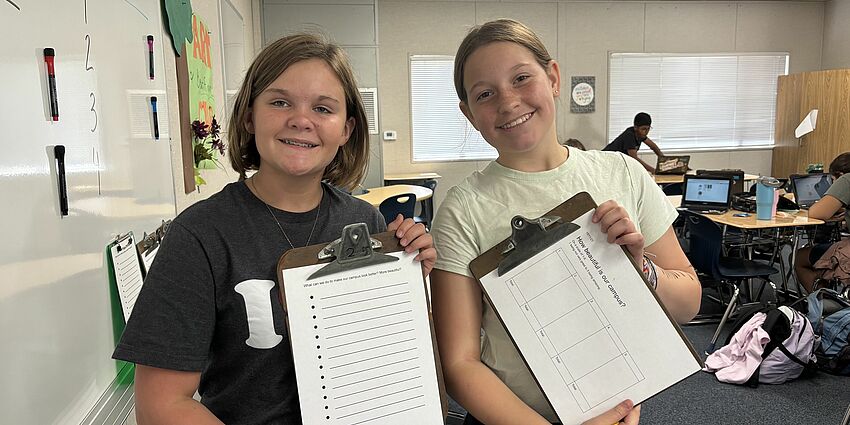 two pre-teen girls standing beside each other, smiling, and holding clipboards showing the forms they used to collect feedback from fellow students about campus beautification