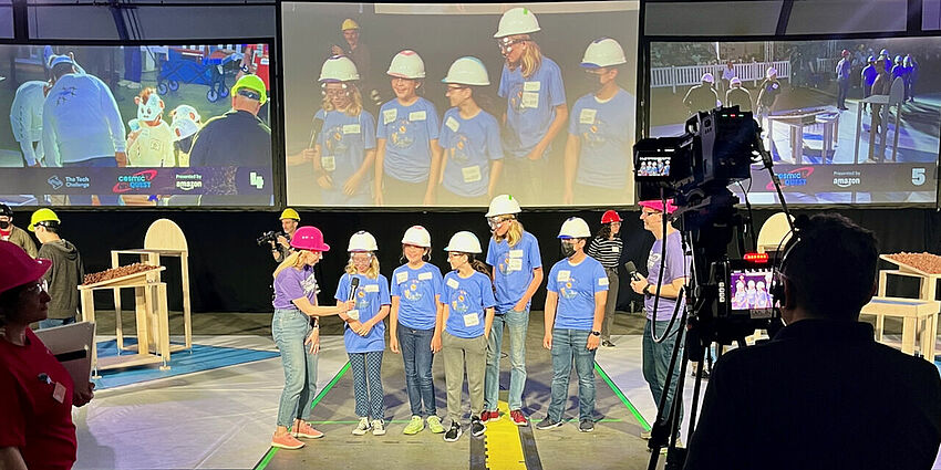 5 students in white hard hats standing in front of cameras as a woman in a pink hard hat holds a microphone  and a man in a pink hard hat listens to what is being said.