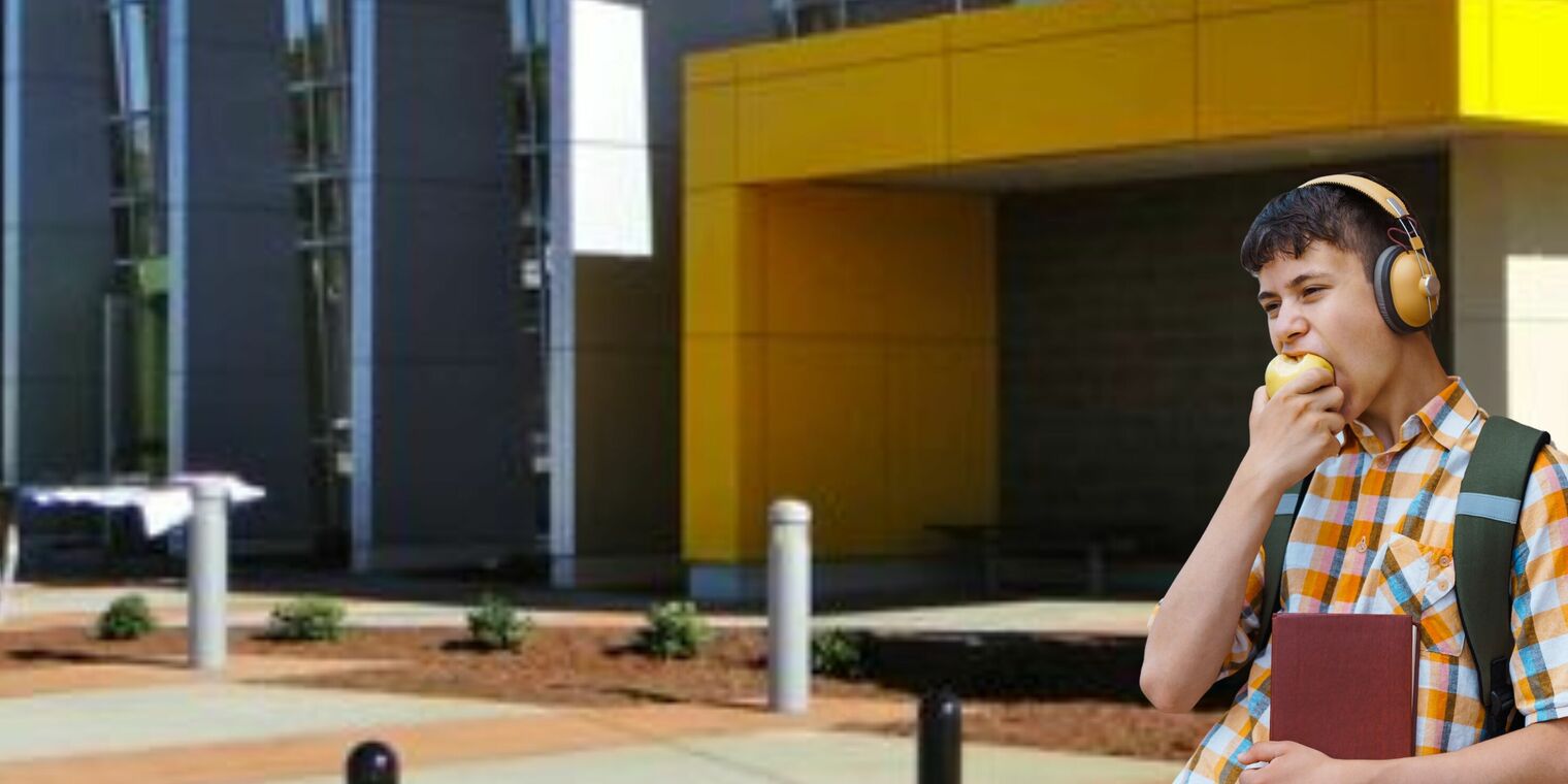 adolescent boy with headphones holding a book and biting into an apple as he stands beside a school building